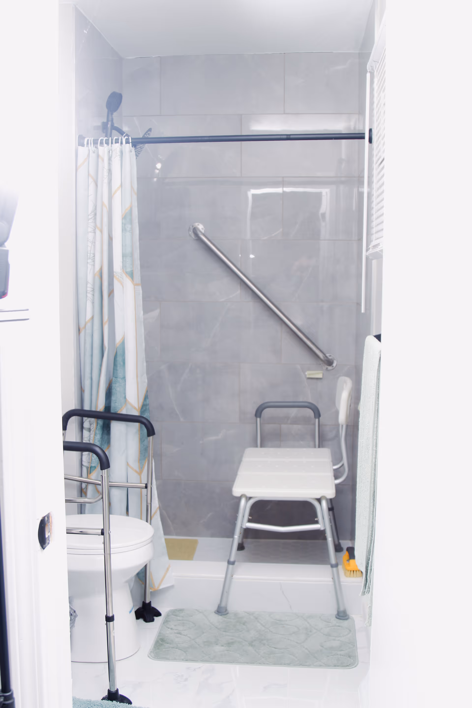 A bathroom with a shower area featuring a shower chair, grab bar, and a shower curtain. There is a toilet with safety rails next to the shower. A towel hangs on the right side, and a bath mat is placed on the floor in front of the shower.