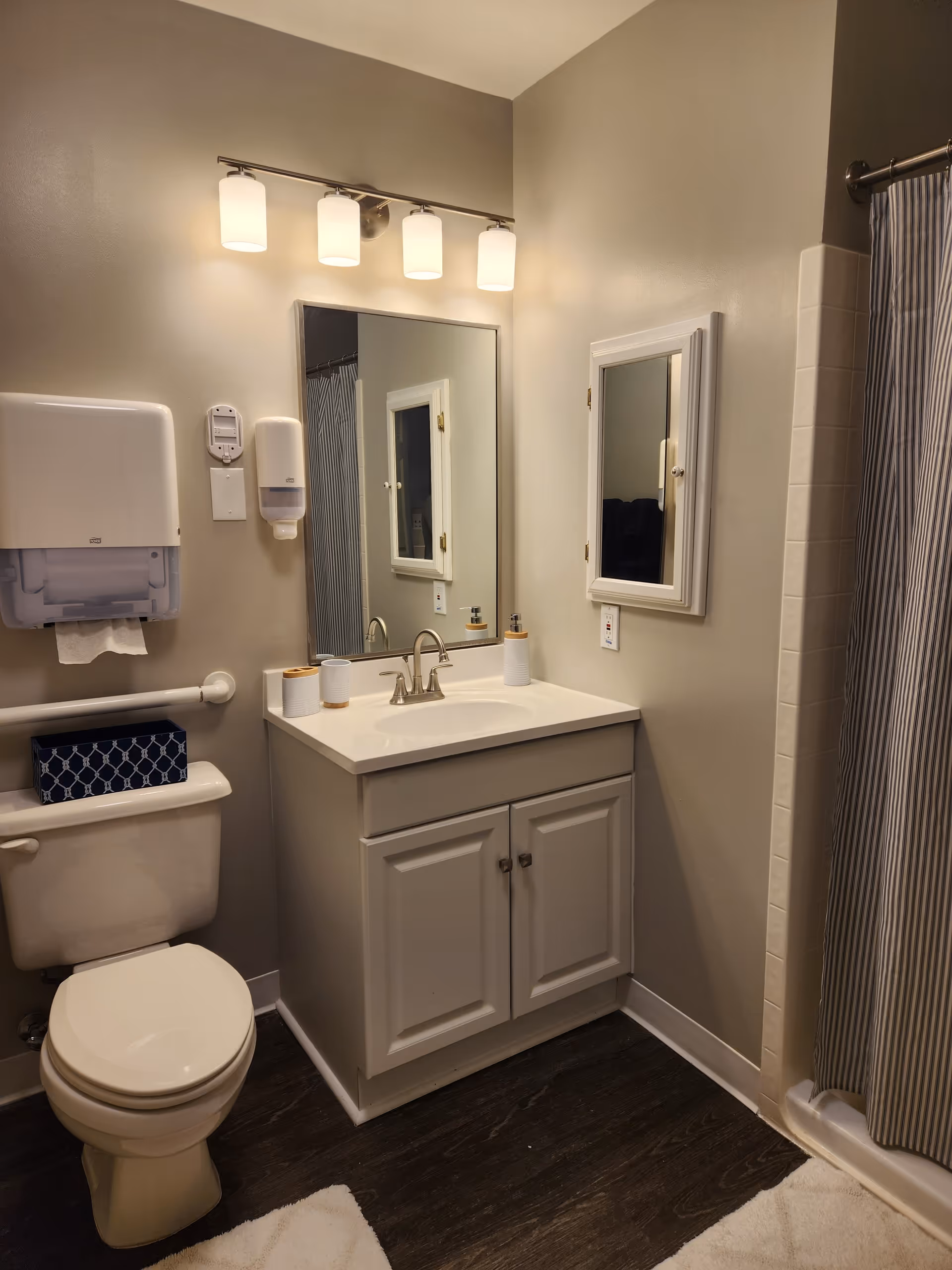 A clean and well-lit bathroom featuring a toilet with a blue patterned tissue box on top, a white vanity with a sink and faucet, a large mirror above the sink, a wall-mounted medicine cabinet with a mirrored door, a shower with a striped curtain, and a four-light fixture above the mirror. The walls are painted beige and the floor has dark wood-like flooring with white bath mats.