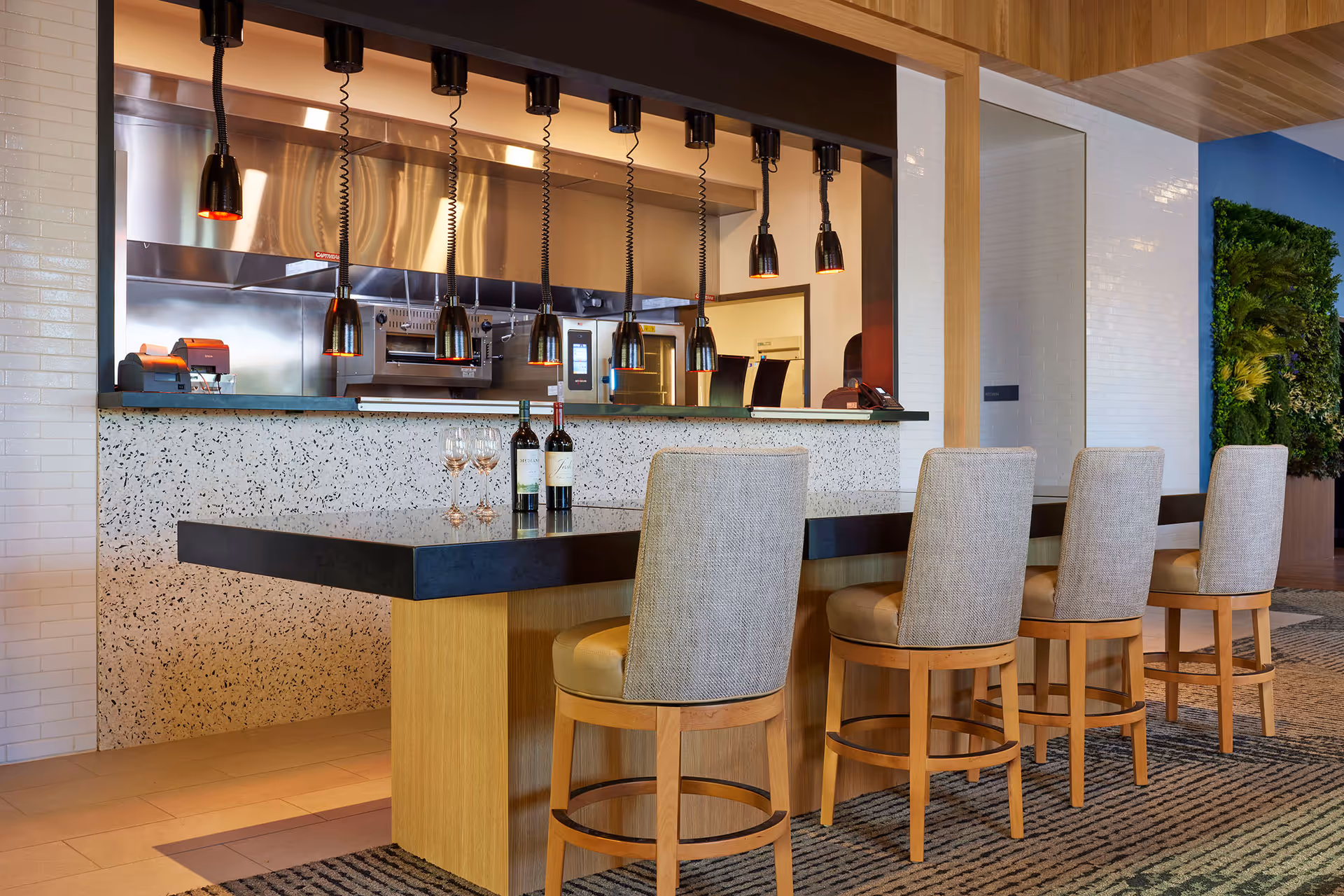 Modern kitchen counter area with four cushioned bar stools, two bottles of wine and two wine glasses on the black countertop. Above the counter are six hanging black pendant heat lamps. The kitchen area behind the counter features stainless steel appliances and a white tiled wall. A green vertical plant wall is visible on the right side.