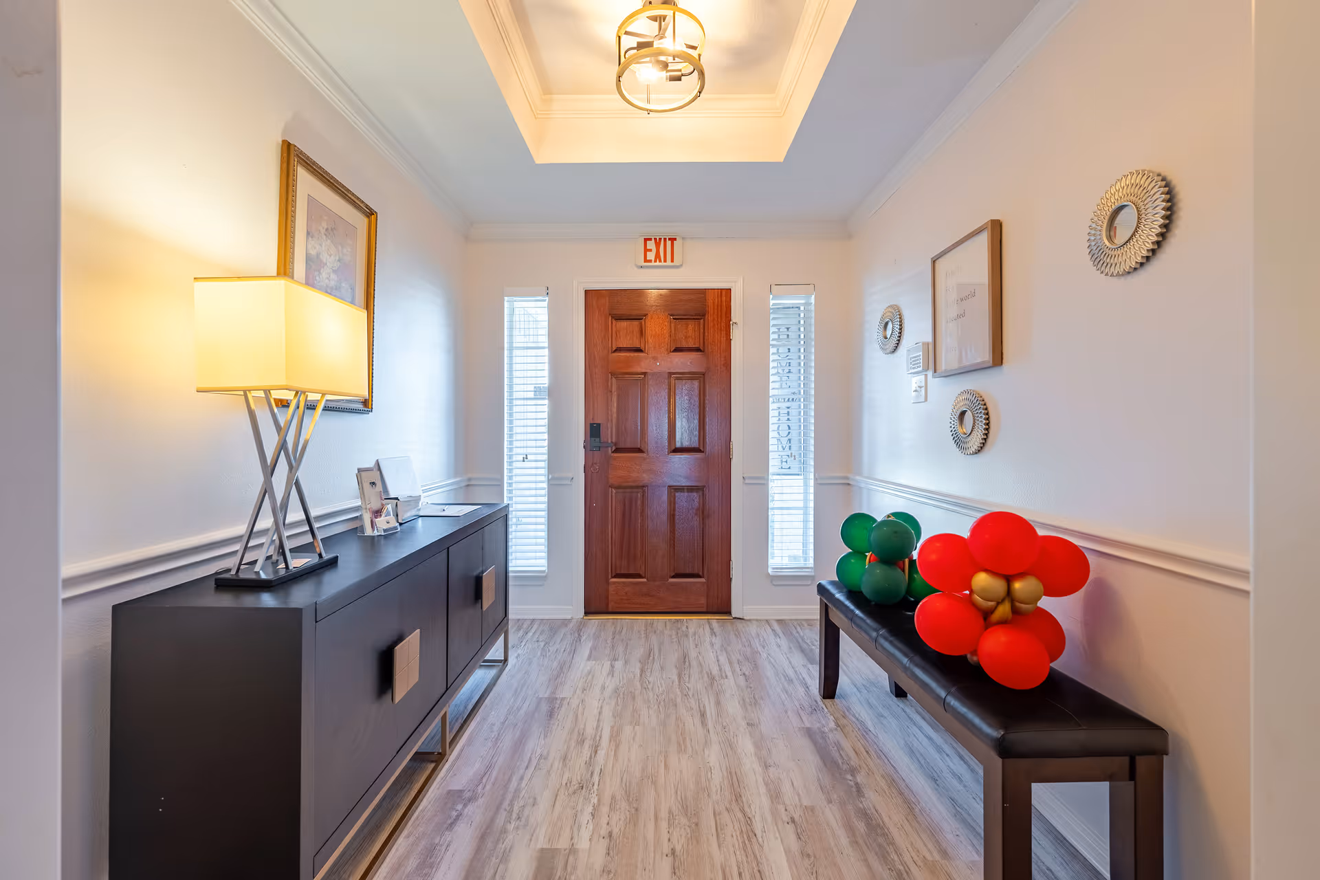 A bright hallway with a wooden front door at the end, flanked by two narrow windows with blinds. On the left side, there is a black console table with a modern lamp and framed pictures. On the right side, a black bench holds balloon decorations shaped like flowers. The walls are decorated with framed art and circular mirrors, and the floor has light wood-style flooring.