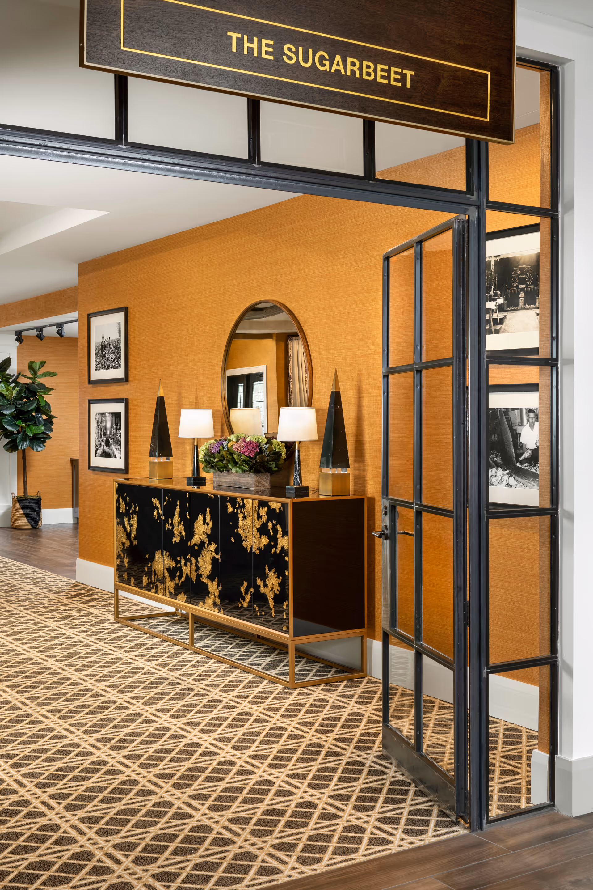 Interior view of a hallway in a senior living facility named The Sugarbeet, featuring a decorative black and gold console table with two lamps and a flower arrangement, an oval mirror on an orange textured wall, framed black and white photographs, a patterned carpet, and a glass-paned door.