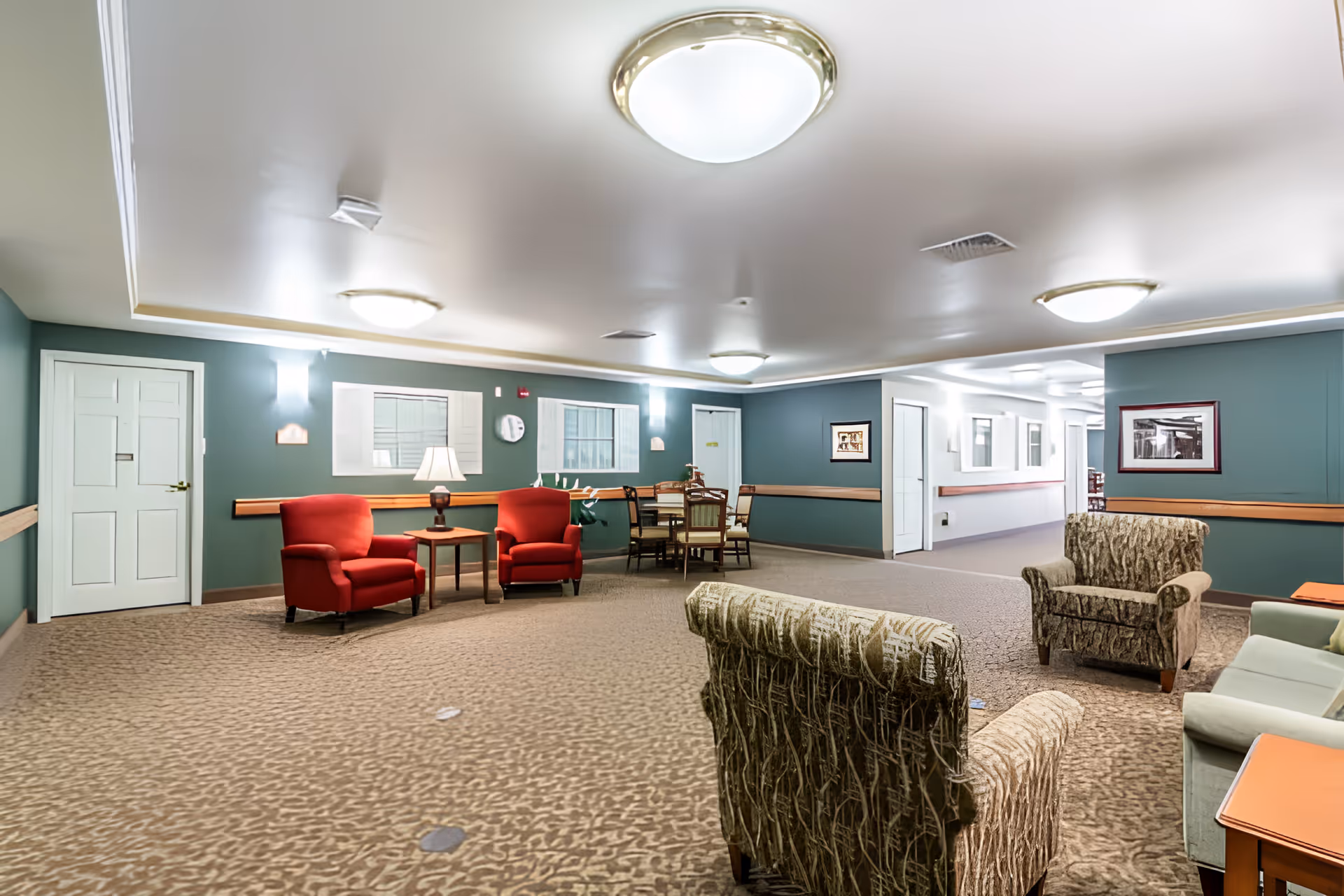 Spacious senior living common room with multiple armchairs and side tables, a dining table in the background, green walls, and ceiling lights.