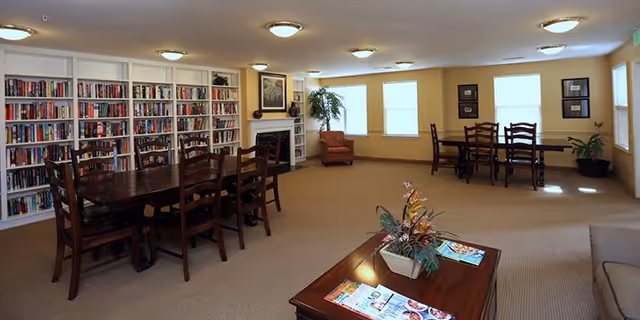 A spacious common room with beige walls and carpet, featuring two wooden tables with chairs, a bookshelf filled with books along one wall, a fireplace with a framed picture above it, a comfortable armchair near windows, and a coffee table with magazines and a floral arrangement.