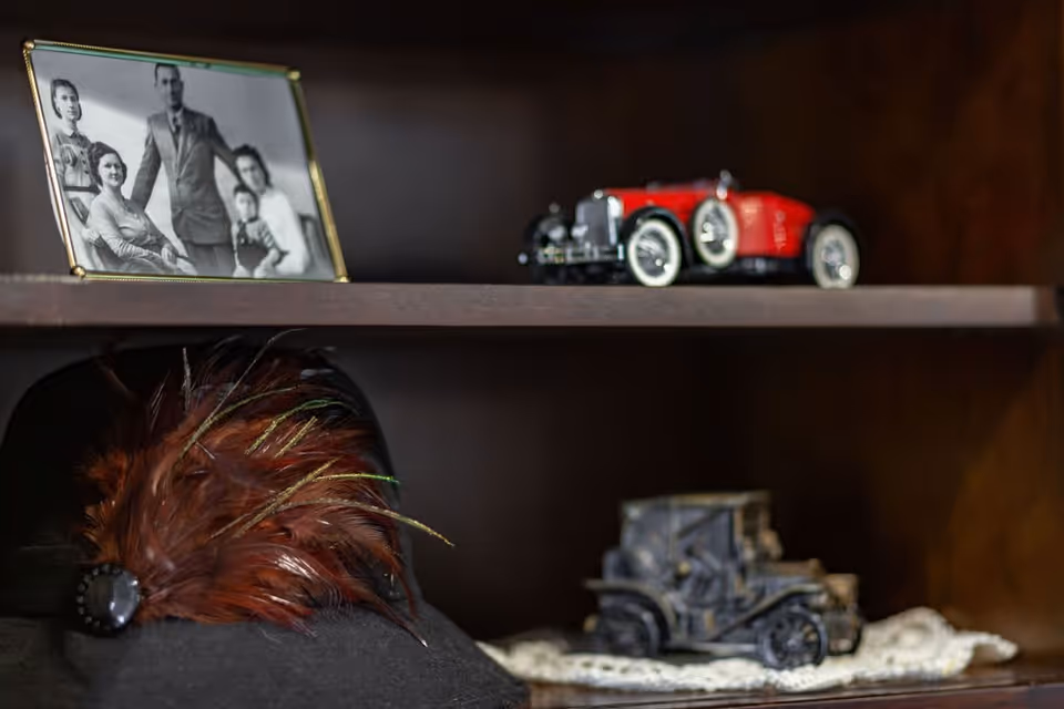 A wooden shelf displaying a black and white framed family photograph, a black hat adorned with brown and green feathers, a red vintage toy car, and a gray vintage toy car placed on a lace doily.