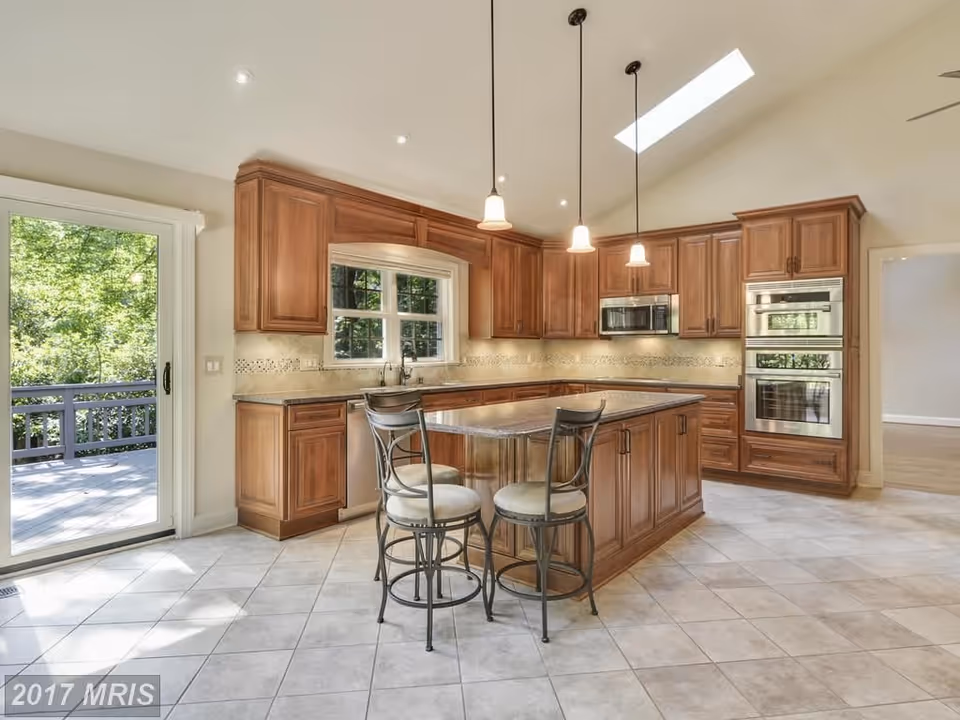 Bright kitchen with wooden cabinets, a central island with two high chairs, tiled floor, pendant lights hanging from the ceiling, a window above the sink, a sliding glass door leading outside, and built-in stainless steel appliances including a microwave and double oven.