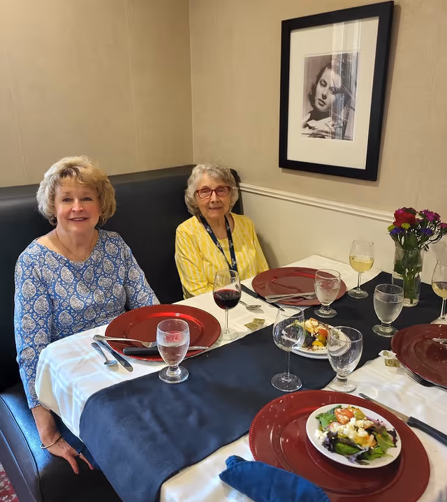 Two elderly women seated at a dining table set with red charger plates, wine glasses, salads, and a vase of flowers in a dining room booth.