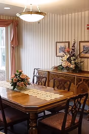 A dining room with a wooden dining table and six chairs. The table has a lace runner and a floral centerpiece. In the background, there is a wooden sideboard with a large floral arrangement and two framed pictures on the wall. A window with pink curtains is partially visible on the left side, and a ceiling light fixture hangs above the table.