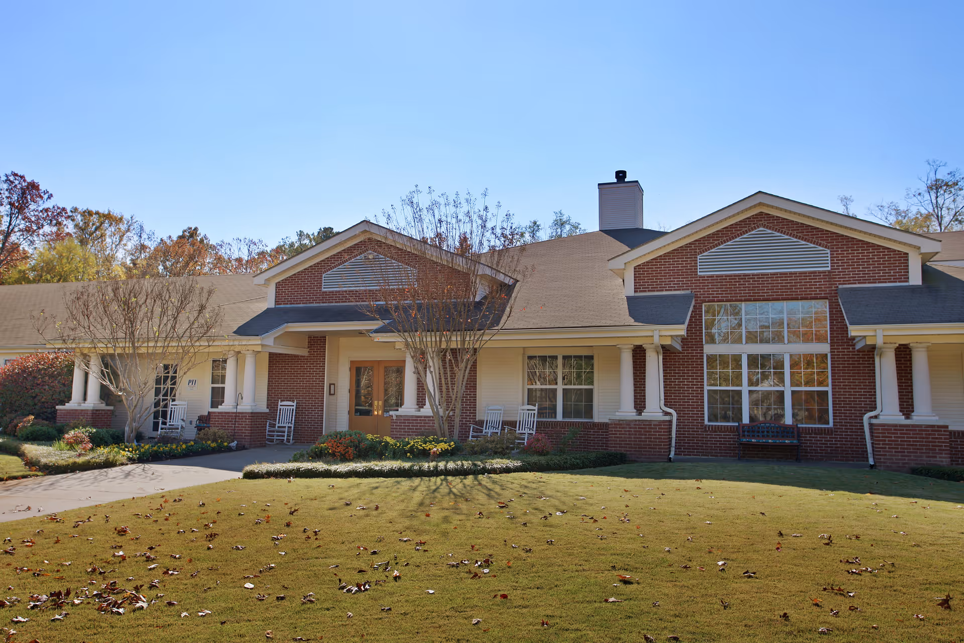 Front exterior view of a single-story brick building with white columns, large windows, and a well-maintained lawn with scattered autumn leaves. There are rocking chairs on the porch and some small trees and shrubs in the landscaping.