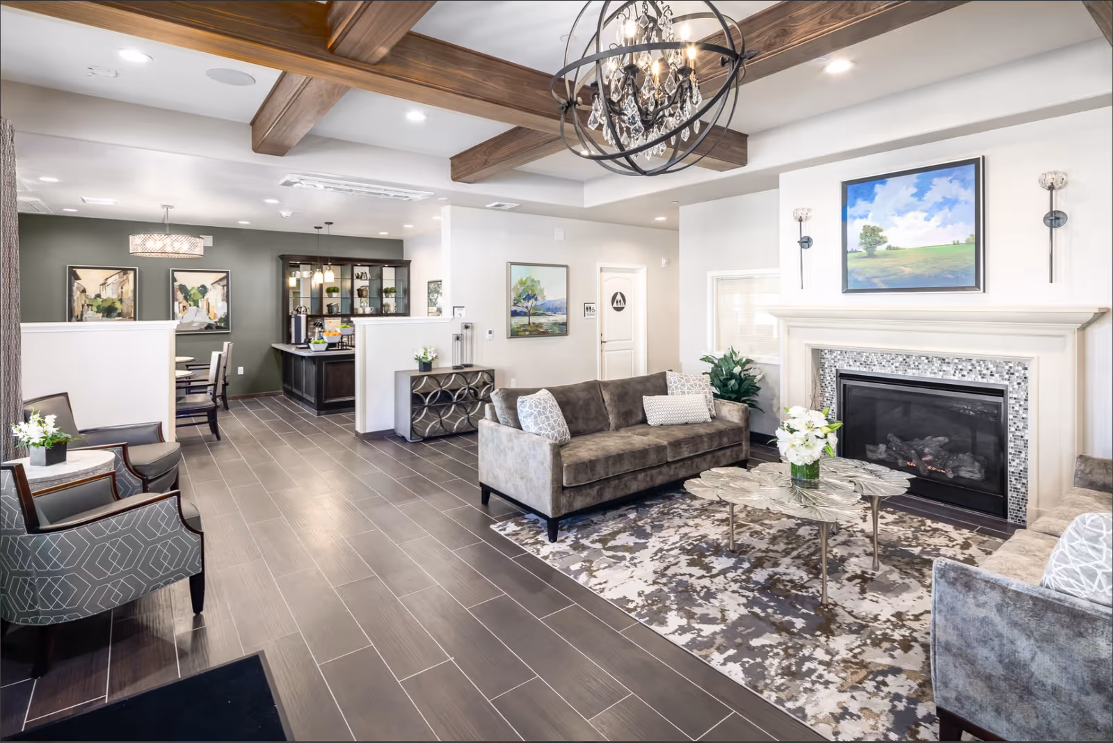 Bright, modern common living room with sofas, a fireplace, a coffee table, chandelier and an adjacent dining/service area.