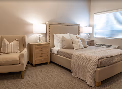 A neatly made bed with beige and white pillows and a beige blanket in a well-lit bedroom. There are two wooden nightstands on either side of the bed, each with a lamp featuring a clear glass base. To the left of the bed is a beige armchair with a striped pillow. A window with closed blinds is on the right wall.