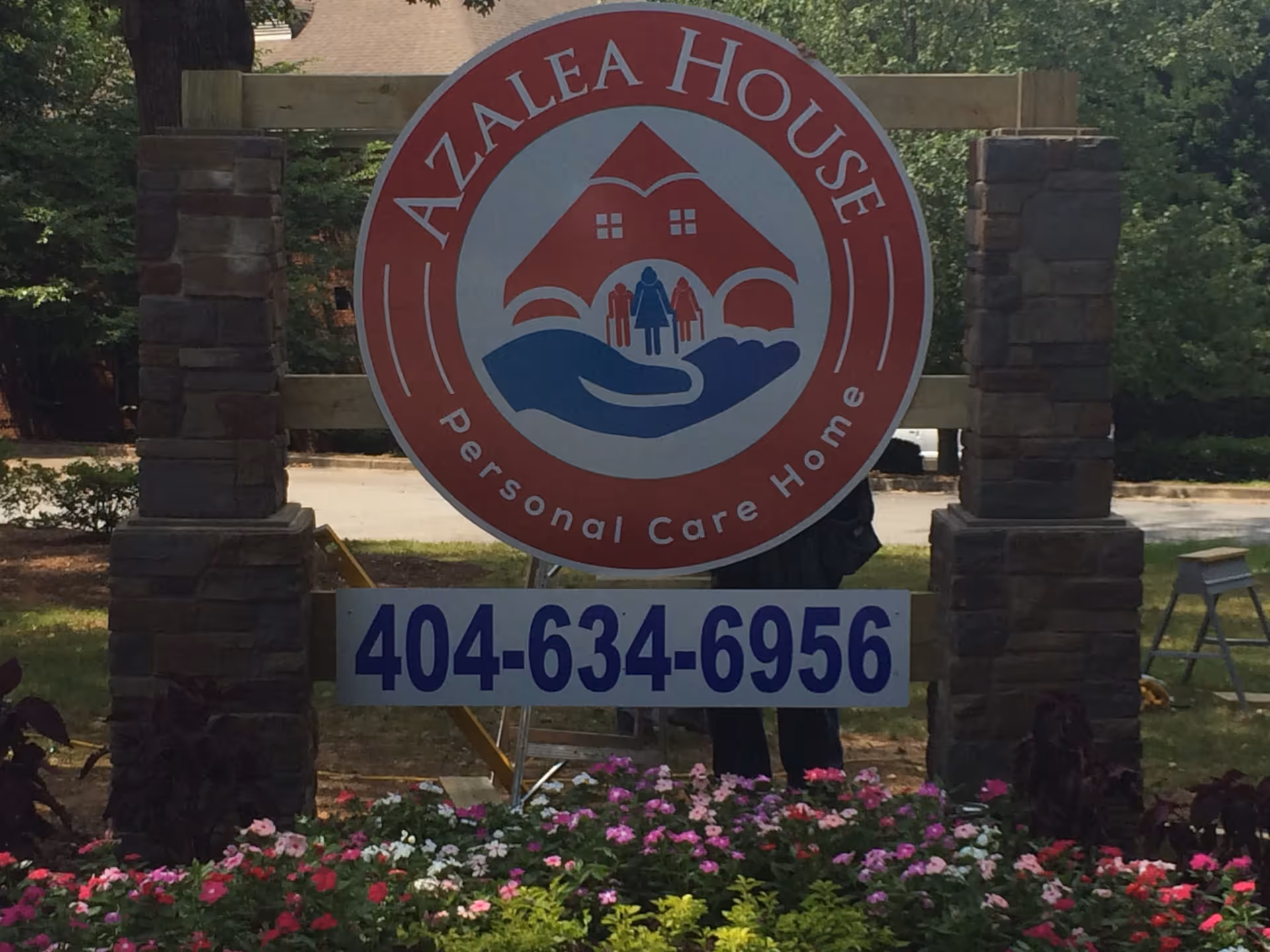 Outdoor sign for Azalea House Personal Care Home mounted on wooden posts with stone bases, surrounded by colorful flowers and greenery.