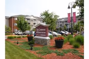 Exterior view of The Commons At The Villages Of Taylor senior living facility showing a landscaped area with a stone sign, flower pots, and a parking lot with cars in front of multi-story residential buildings.