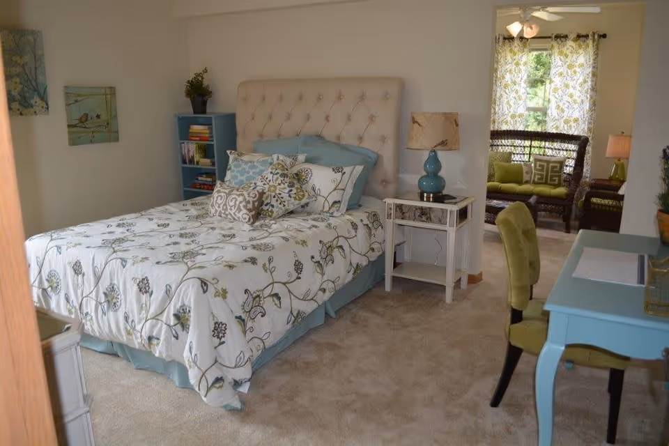 A cozy bedroom with a bed featuring a tufted headboard and floral bedding. There is a blue bookshelf with books and a plant, a white nightstand with a blue lamp, and a green upholstered chair at a blue desk. In the background, a sitting area with wicker furniture and green cushions is visible near a window with floral curtains.