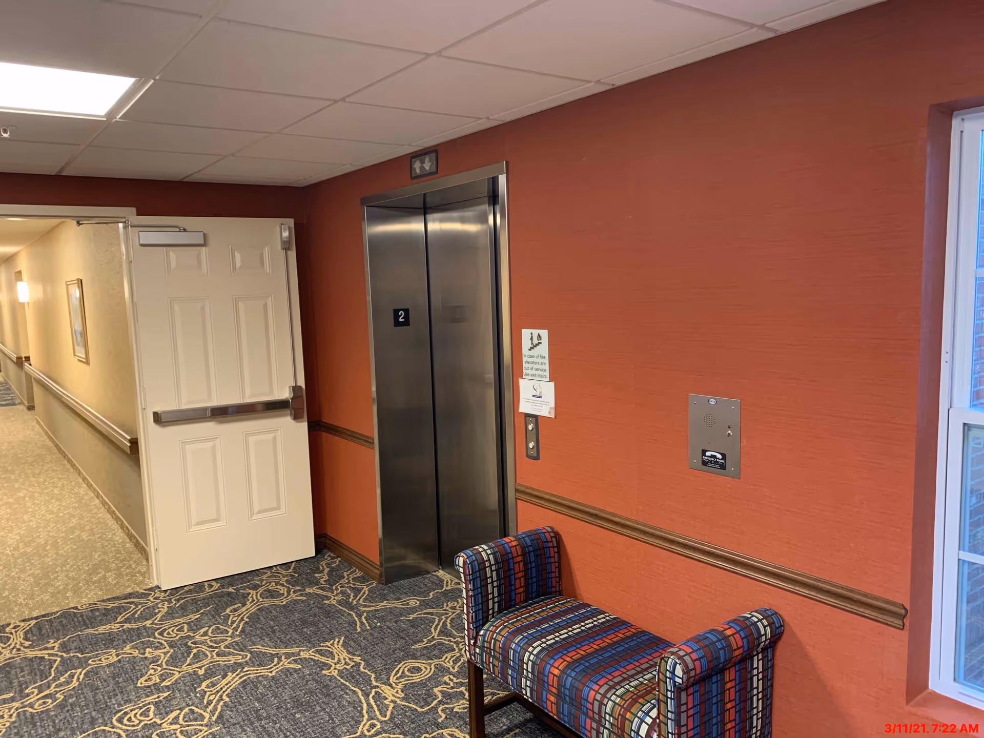 Interior hallway of Creek Valley Retirement Community showing a closed elevator with stainless steel doors marked with the number 2, a colorful patterned bench against a red wall, an open white door leading to a corridor with beige walls and carpeted floor, and a window on the right side.
