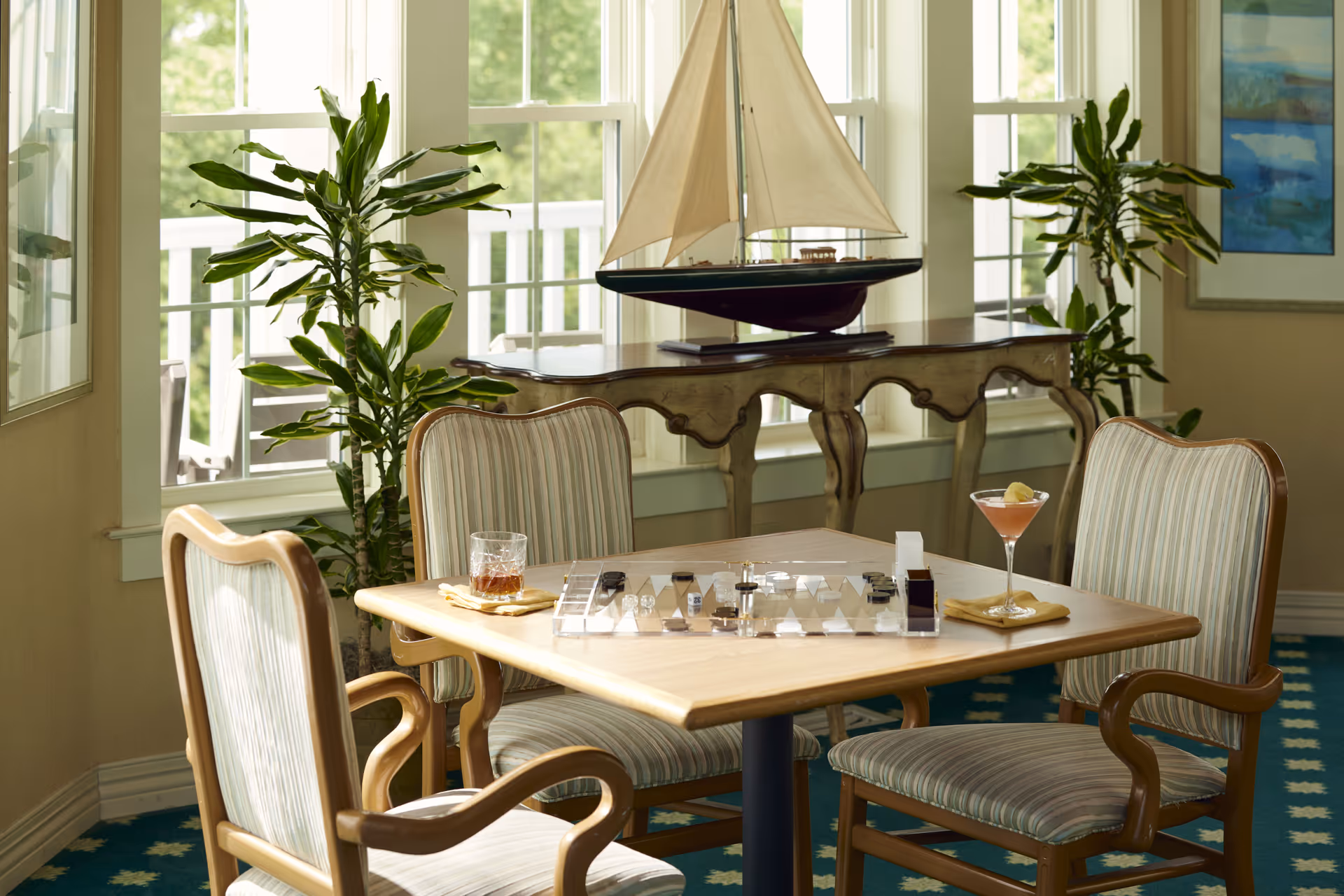 Sunlit seating area with a wooden table set for drinks and a board game, surrounded by upholstered chairs and a model sailboat on a console by the window.