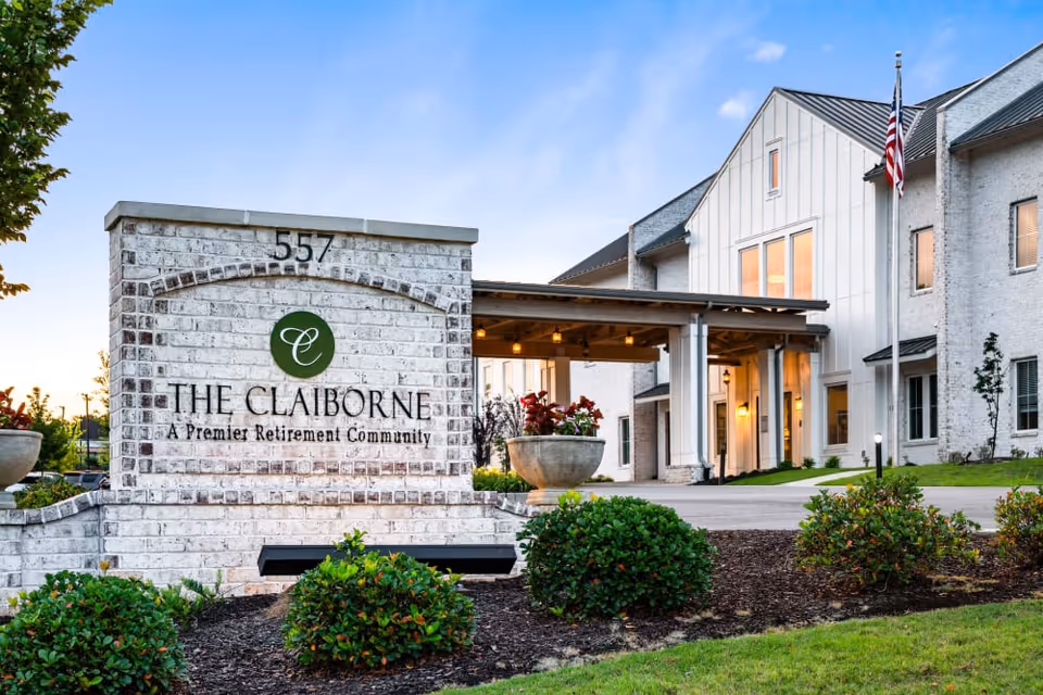 Front entrance and sign for The Claiborne retirement community with a covered portico, American flag, and landscaped grounds.