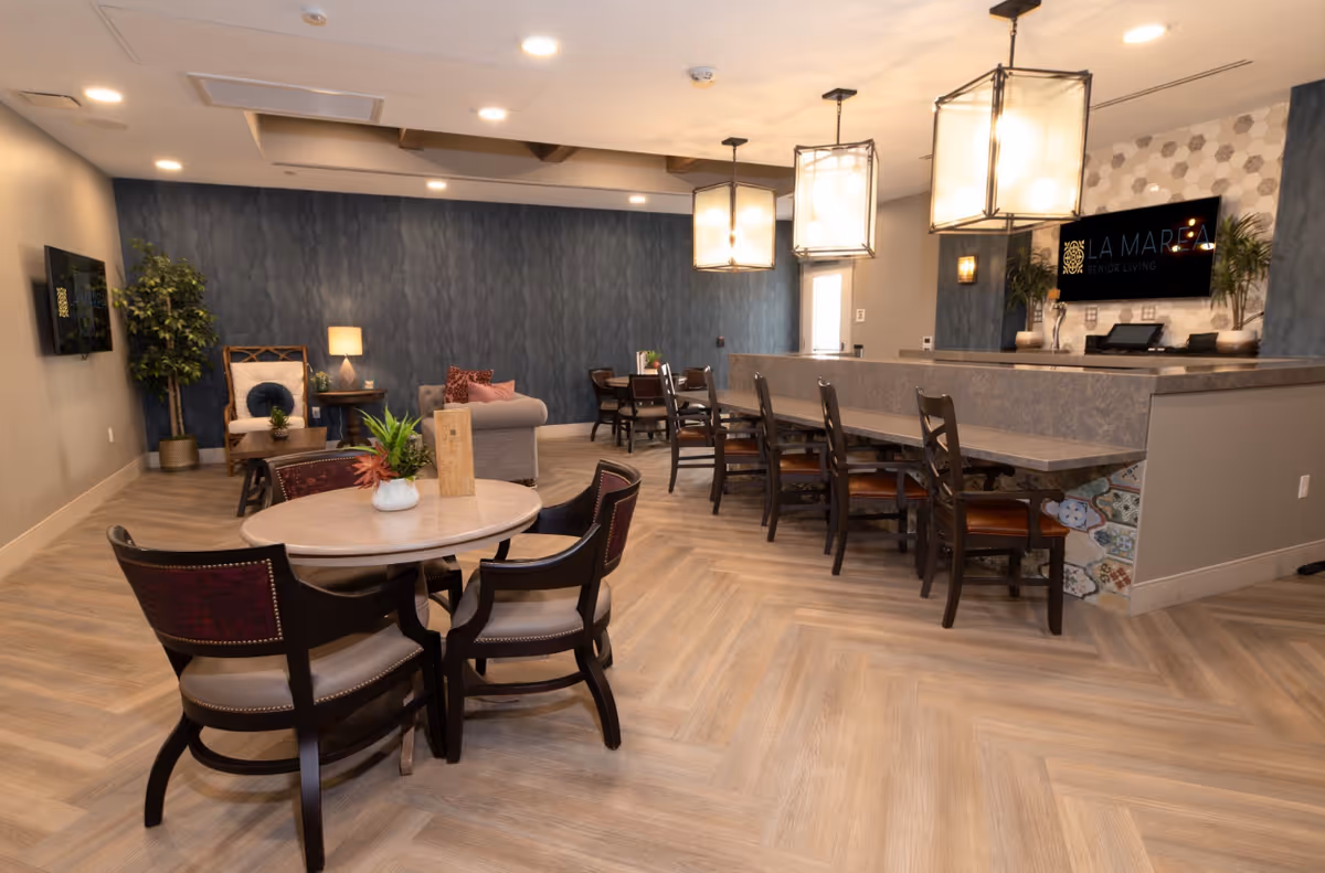 A modern senior living common area with a round table and four chairs in the foreground, a long counter with several chairs, and a cozy seating area with a sofa, armchair, and side table against a dark blue accent wall. The room features wood-patterned flooring, pendant lights hanging from the ceiling, and a TV screen displaying the La Marea Senior Living logo.
