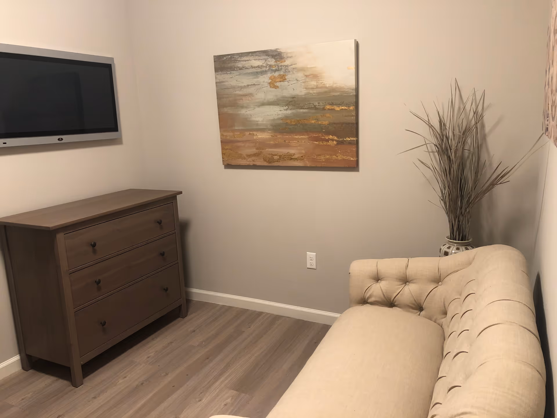 A small room with a beige tufted sofa on the right, a wooden dresser with three drawers on the left, a flat-screen TV mounted on the wall above the dresser, an abstract painting with earthy tones hanging on the wall, and a tall vase with decorative dried grass in the corner.