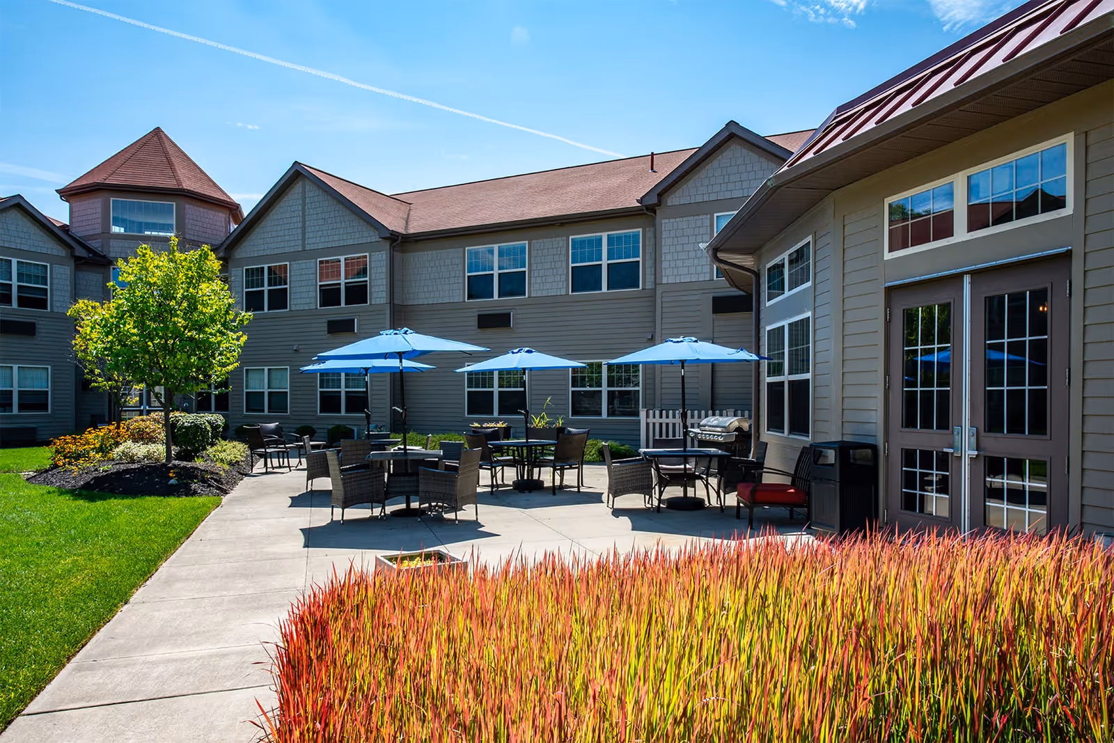Outdoor patio area at Beachwood Commons by New Perspective featuring several tables with blue umbrellas, surrounded by chairs. The patio is adjacent to a two-story building with multiple windows and a red roof. There is a well-maintained lawn, a small tree, and ornamental grasses in the foreground under a clear blue sky.