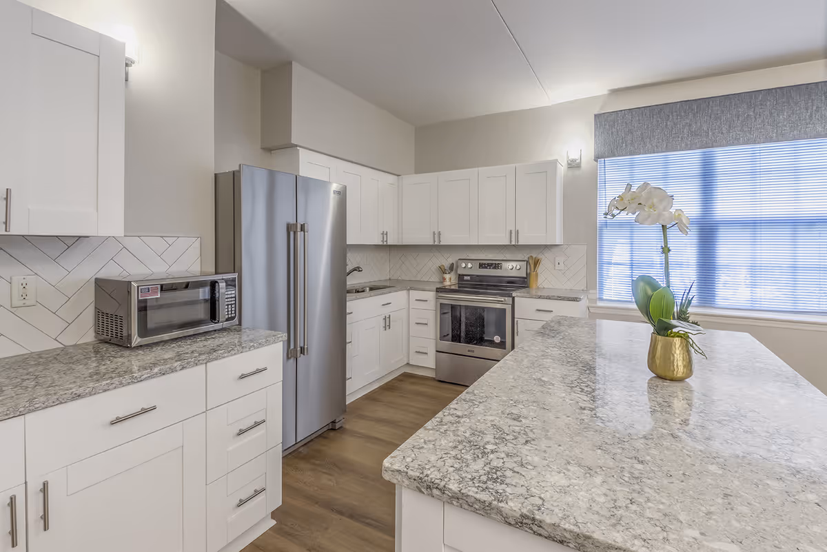 Modern kitchen with white cabinets, a stainless steel refrigerator and stove, a microwave on the counter, and a large marble countertop island with a small potted orchid plant. A window with blinds is visible in the background.
