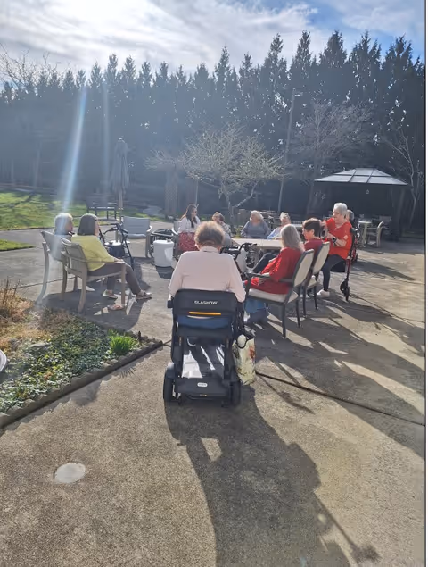 A group of elderly people sitting in a circle outdoors on a sunny day, surrounded by trees and garden area. Some are seated on chairs while one person is in a wheelchair. There is a gazebo in the background and the setting appears to be a communal outdoor space at a senior living facility.