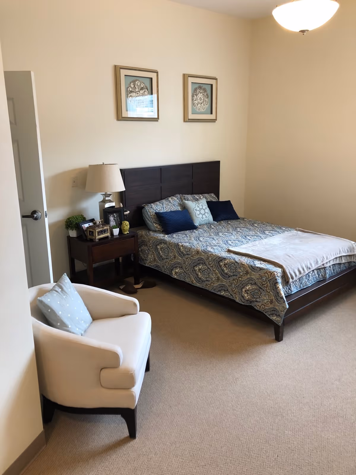 A bedroom with a double bed featuring a patterned blue and beige bedspread and multiple pillows. There is a dark wooden nightstand with a lamp, framed photos, and small decorative items next to the bed. A white upholstered armchair with a light blue pillow is positioned near the door. Two framed artworks hang on the wall above the bed. The room has beige carpeting and cream-colored walls.