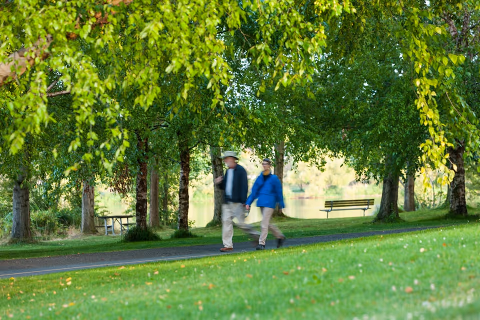 Two people walking on a paved path in a green park area with trees, grass, a bench, and a picnic table visible in the background.