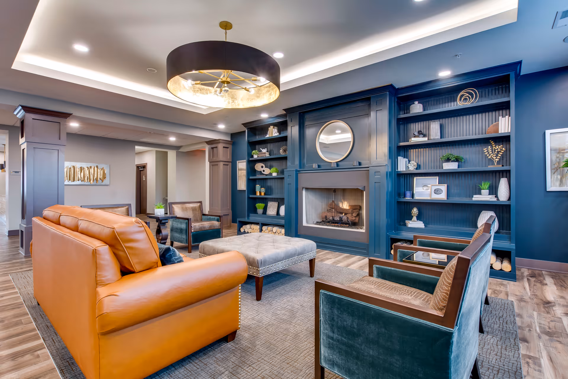 Stylish lounge with leather sofa, velvet armchairs, central ottoman, a built-in bookshelf and fireplace under a decorative ceiling fixture.