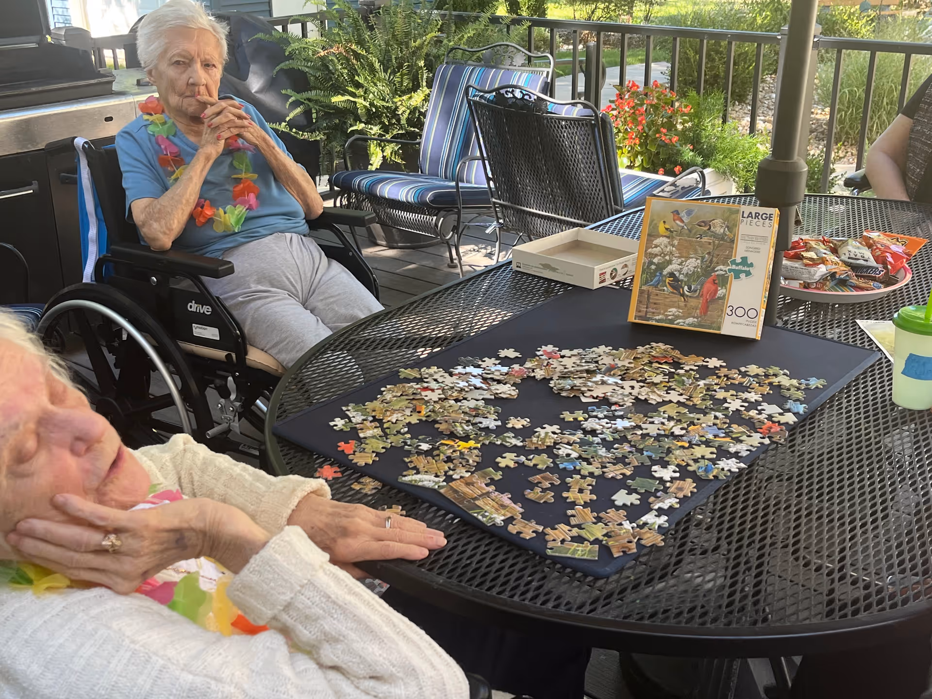 Two elderly women sitting at a black metal outdoor table with a partially completed jigsaw puzzle spread out on a black mat. One woman is in a wheelchair wearing a blue shirt and a colorful lei, while the other woman is resting her head on her hand wearing a white sweater and a colorful lei. The table also has a puzzle box, a plate of snacks, and a green cup. The setting appears to be a patio with outdoor furniture and plants in the background.