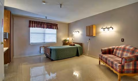 A simple, clean room with a single hospital-style bed covered with a green blanket, a plaid upholstered couch, a window with blinds and a valance, wall-mounted lights, and a small wooden cabinet on the wall.