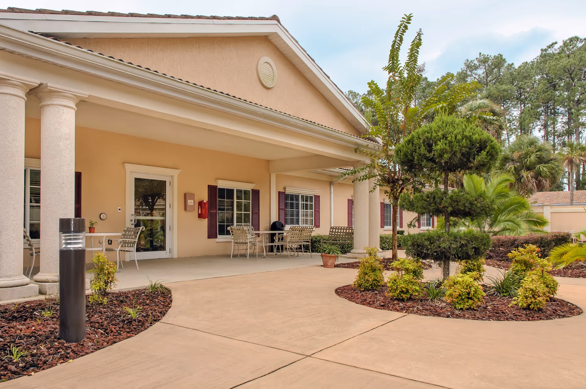 Covered front entrance of a beige senior living building with columns, outdoor seating, and landscaped walkways.