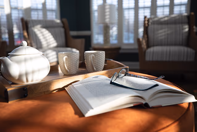 A sunlit living room scene with an open book and reading glasses on a table, and a tray holding a teapot and two cups in front of upholstered armchairs and windows.