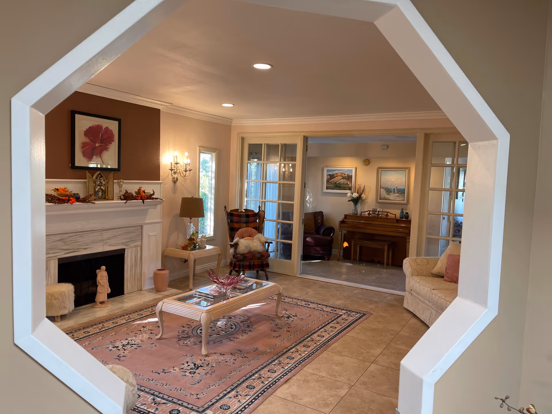 View through a white octagonal interior window frame into a cozy living room with a fireplace decorated with autumn-themed items and a framed flower picture above it. The room has a pink patterned rug, a glass coffee table, a plaid armchair with a white throw, a side table with a lamp, and a beige sofa with a pink pillow. Beyond the living room, there is another room with a piano, two framed landscape paintings, and a brown leather chair.