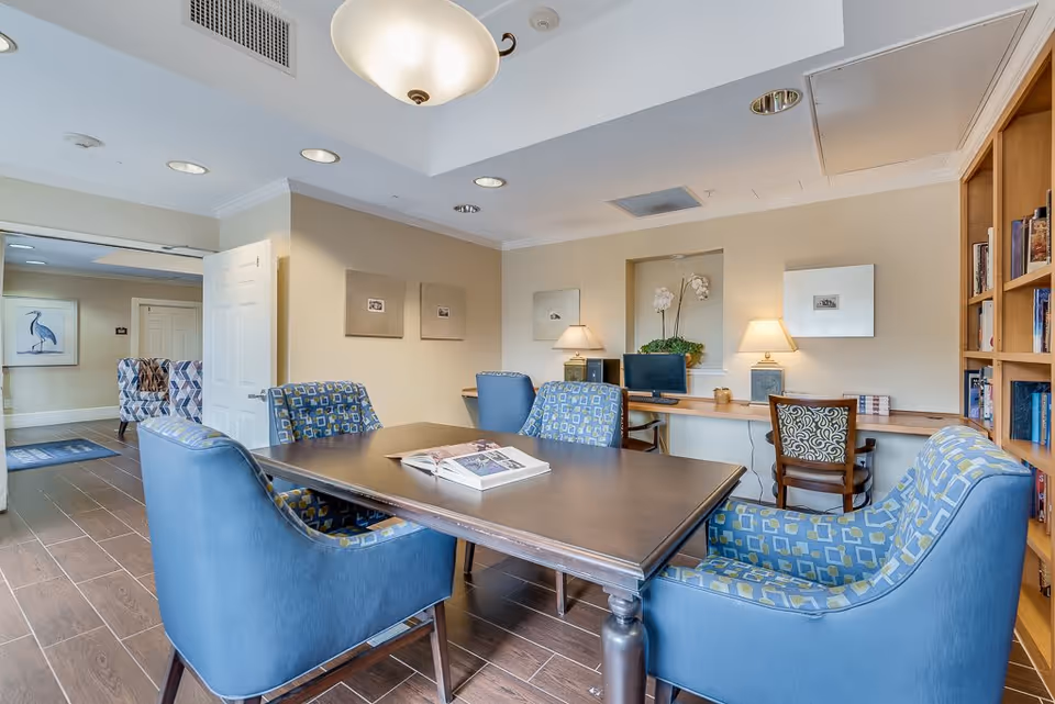 A cozy interior room with a wooden table surrounded by six blue patterned armchairs. There are two computer workstations with chairs and lamps against the far wall, a bookshelf filled with books on the right, and a doorway leading to another room with additional seating and a framed bird picture on the wall.