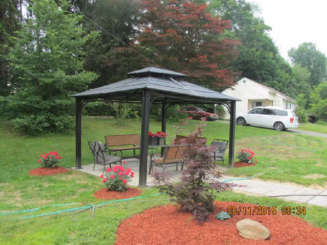 Outdoor garden area with a black metal gazebo containing benches and chairs, surrounded by green grass, red flowering plants, and trees. A white vehicle and a small white building are visible in the background.