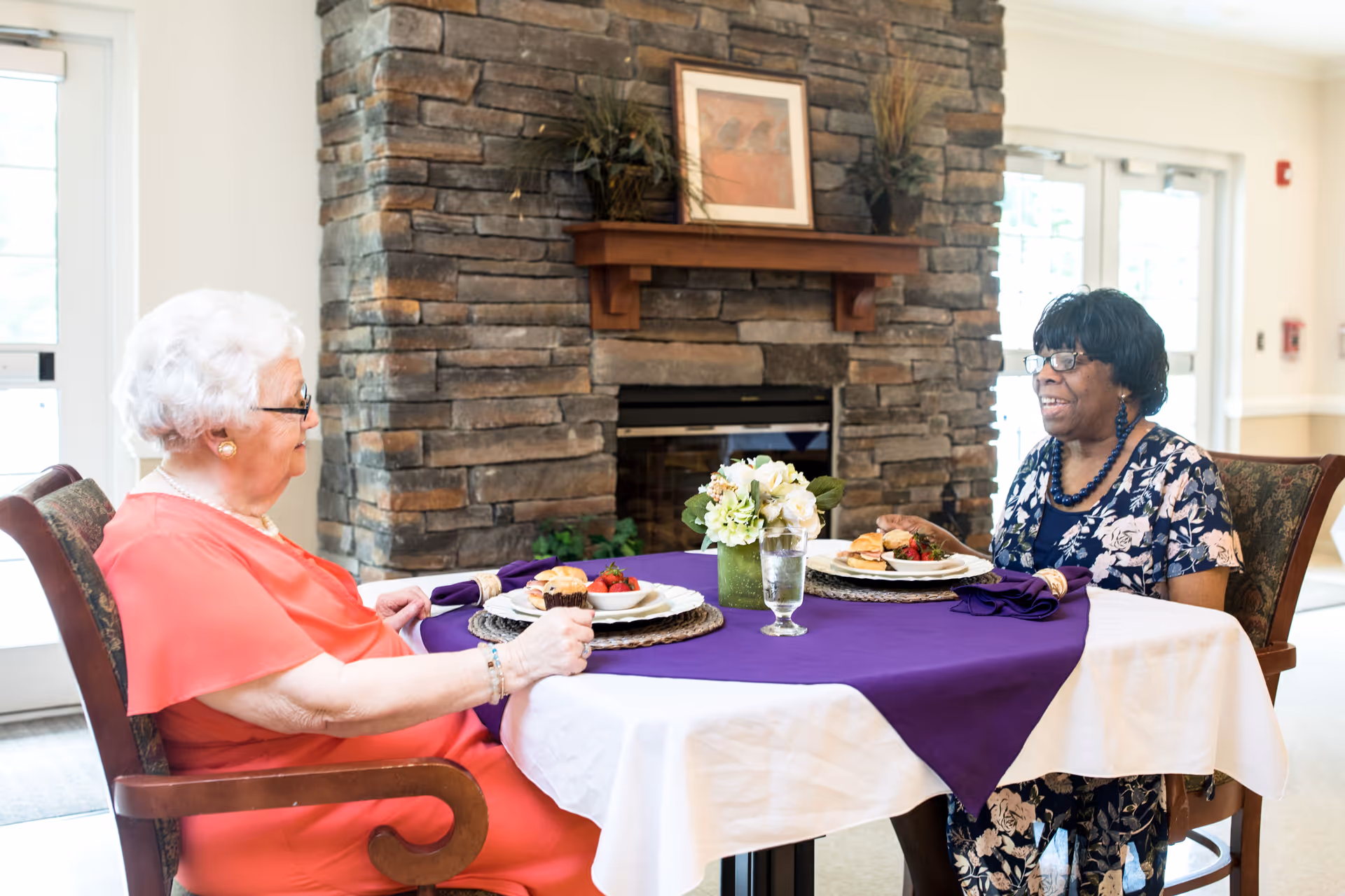 Two elderly women sitting at a round dining table covered with a white tablecloth and a purple runner, enjoying a meal together in a room with a stone fireplace and large windows.