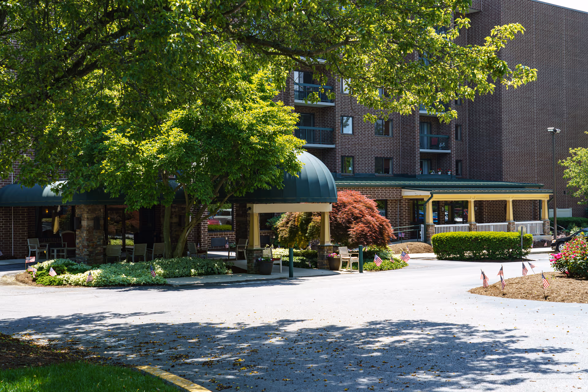 Front entrance of a brick senior living building with a green canopy, trees, landscaping, and small American flags along the driveway.