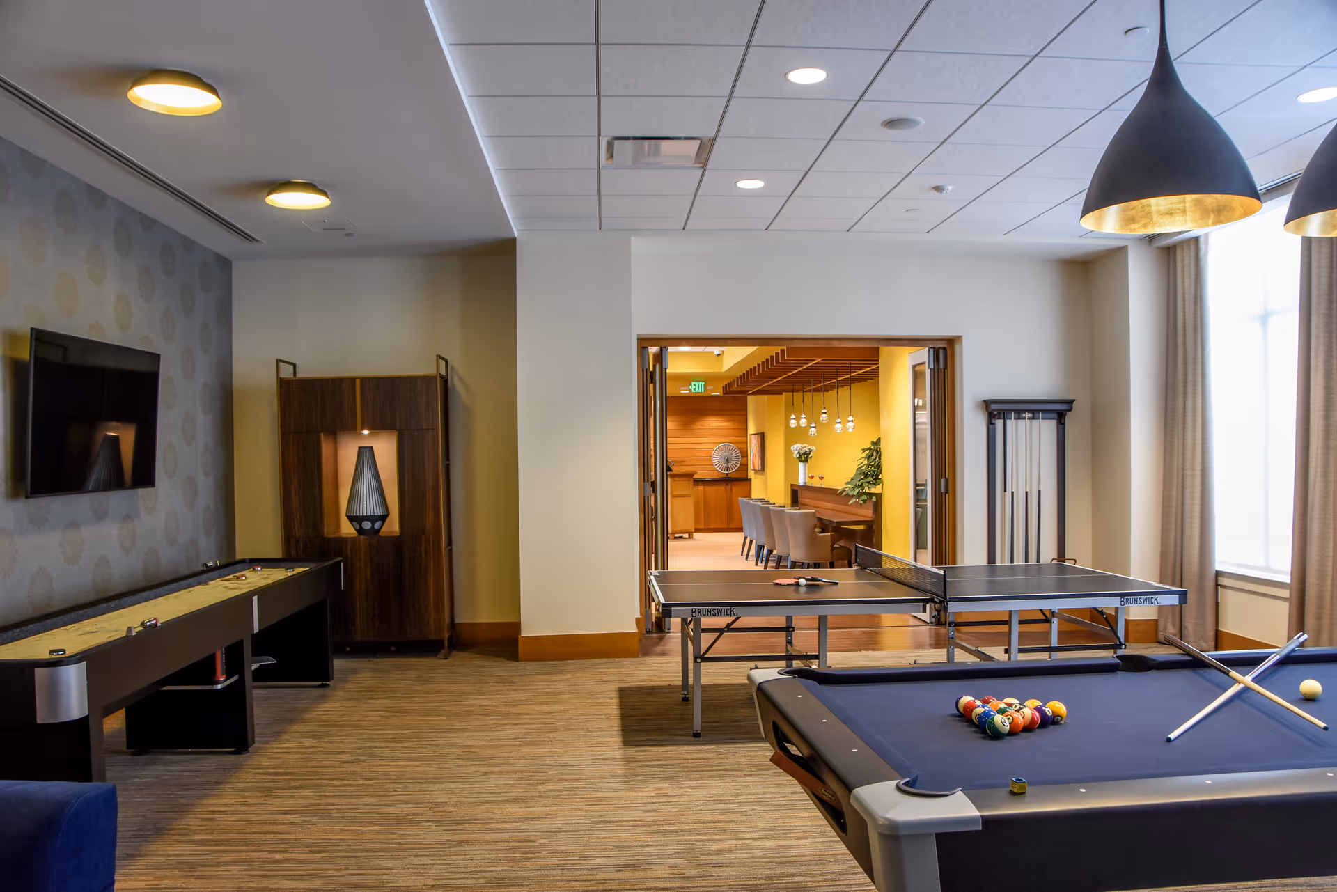 A recreational room with a pool table, a ping pong table, and a shuffleboard table. The room has modern lighting fixtures, a wall-mounted TV, and a cabinet with a decorative lamp. In the background, there is an adjoining room with a long table and chairs, decorated with plants and pendant lights.