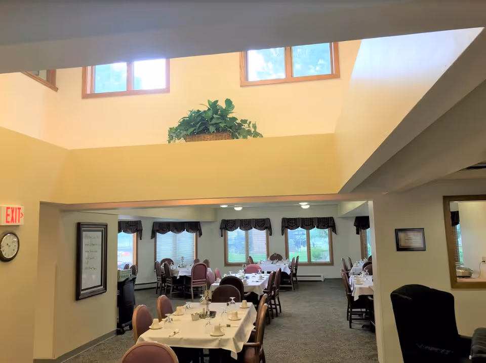 Interior view of a senior community dining room with multiple tables covered in white tablecloths, set with cups and utensils. The room has several windows with valances, a clock on the wall, and a potted plant on a ledge above the entrance.