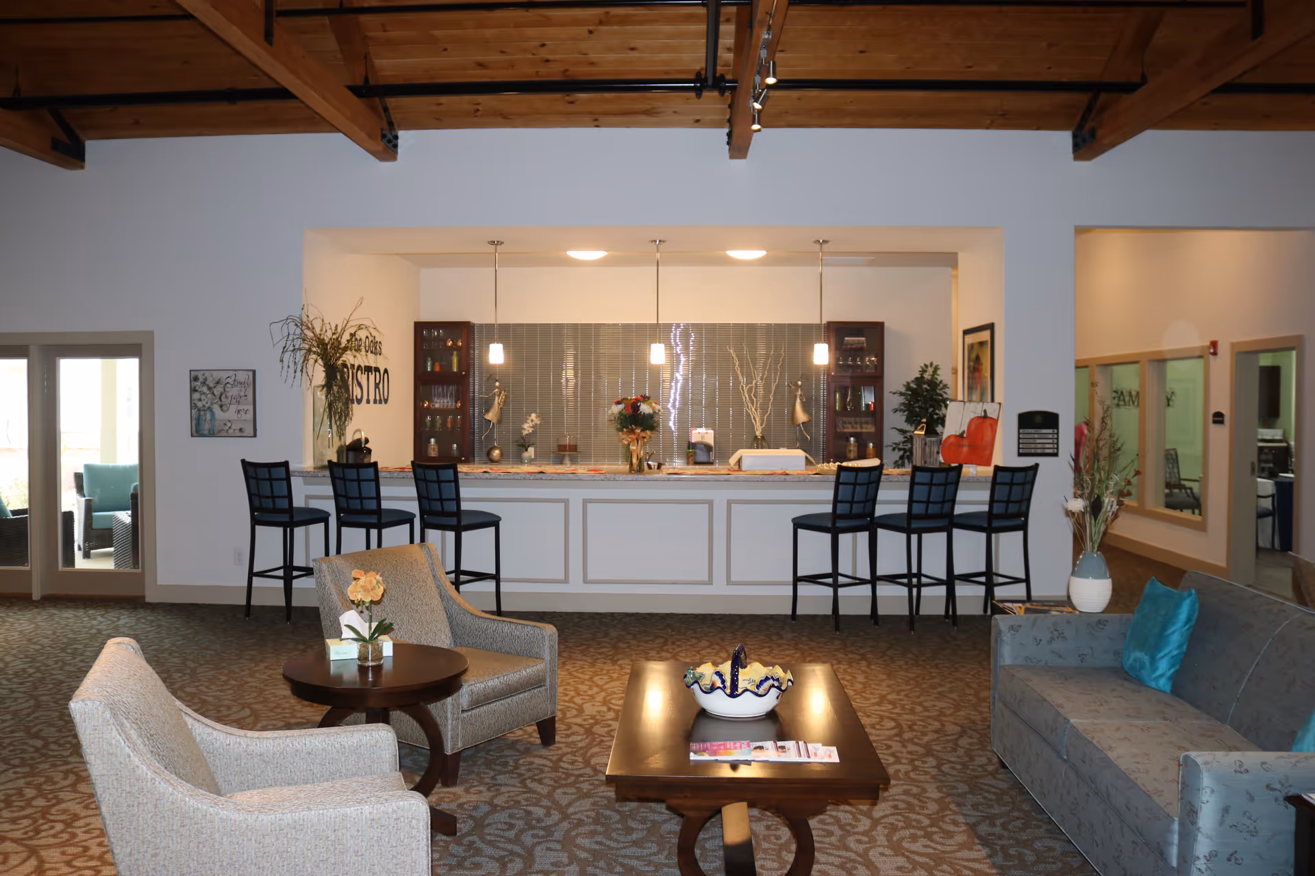 Open common lounge with seating, a coffee table, and a bistro-style counter with bar stools beneath exposed wooden ceiling beams.