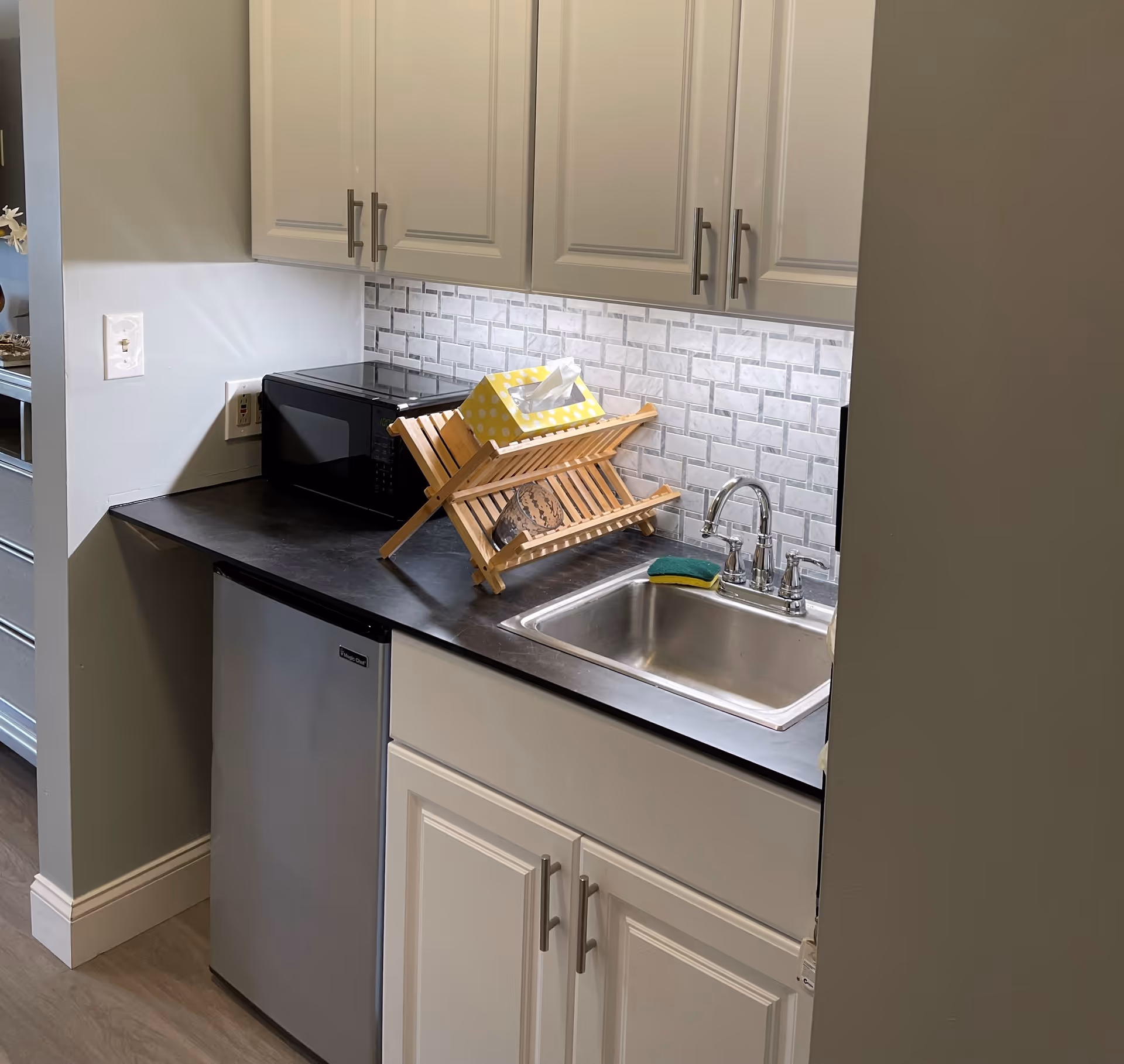 A small kitchenette area with white cabinets, a black countertop, a stainless steel sink with a chrome faucet, a black microwave, a wooden dish rack holding a yellow tissue box and a cup, and a mini refrigerator below the counter. The backsplash features a white and gray tile pattern.