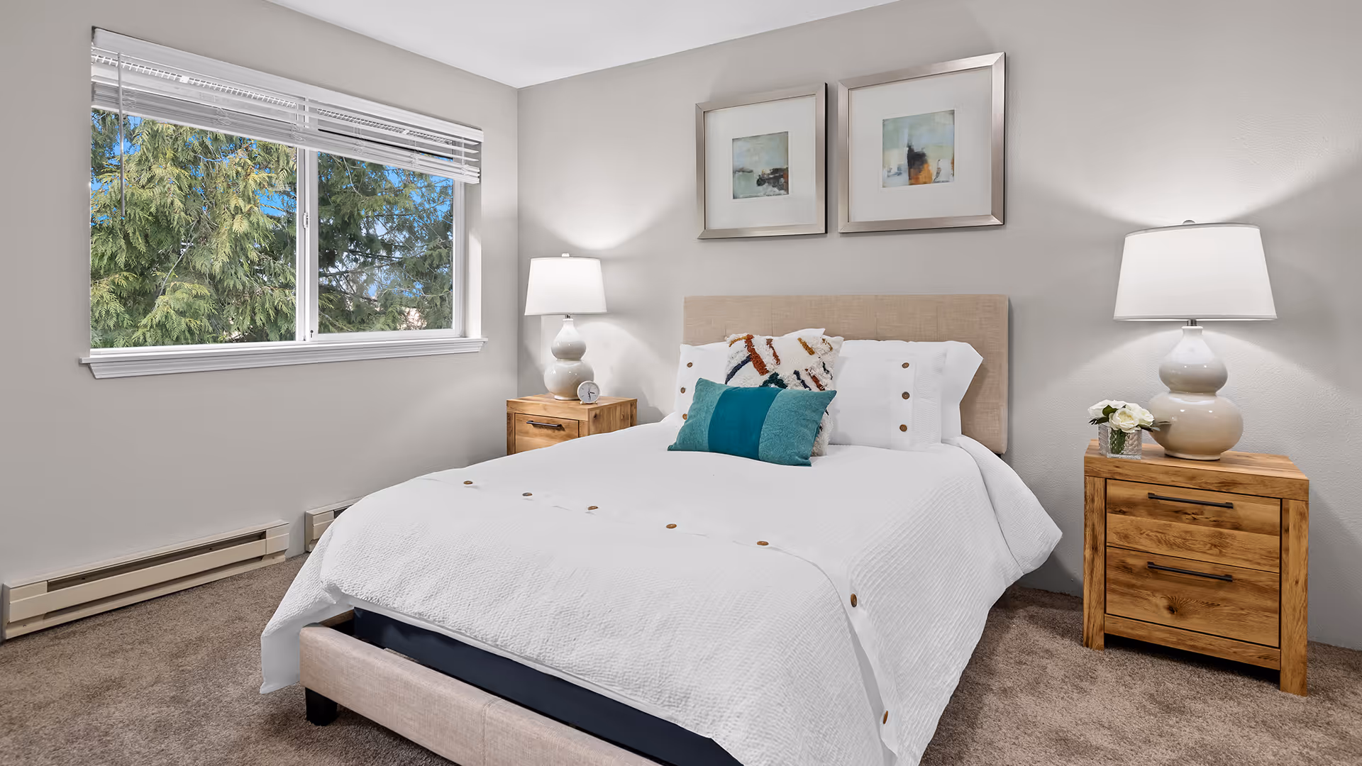 A neatly made bed with white bedding and decorative pillows in a bedroom. The bed has a beige upholstered headboard and is flanked by two wooden nightstands, each with a white lamp. Above the bed are two framed abstract paintings. A window on the left side shows green trees outside. The room has beige carpet and light gray walls.