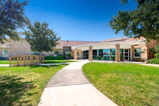Front exterior entrance of a senior living facility with a curved concrete walkway, grassy lawns, trees, and a low brick building with large windows.