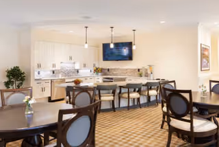 Communal dining area with round tables and chairs facing a modern open kitchen with bar seating and a wall-mounted TV.