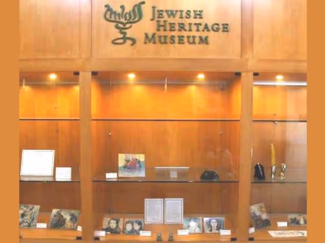 Wooden display case with glass shelves showcasing various framed photographs and documents, with a sign above reading 'Jewish Heritage Museum'.