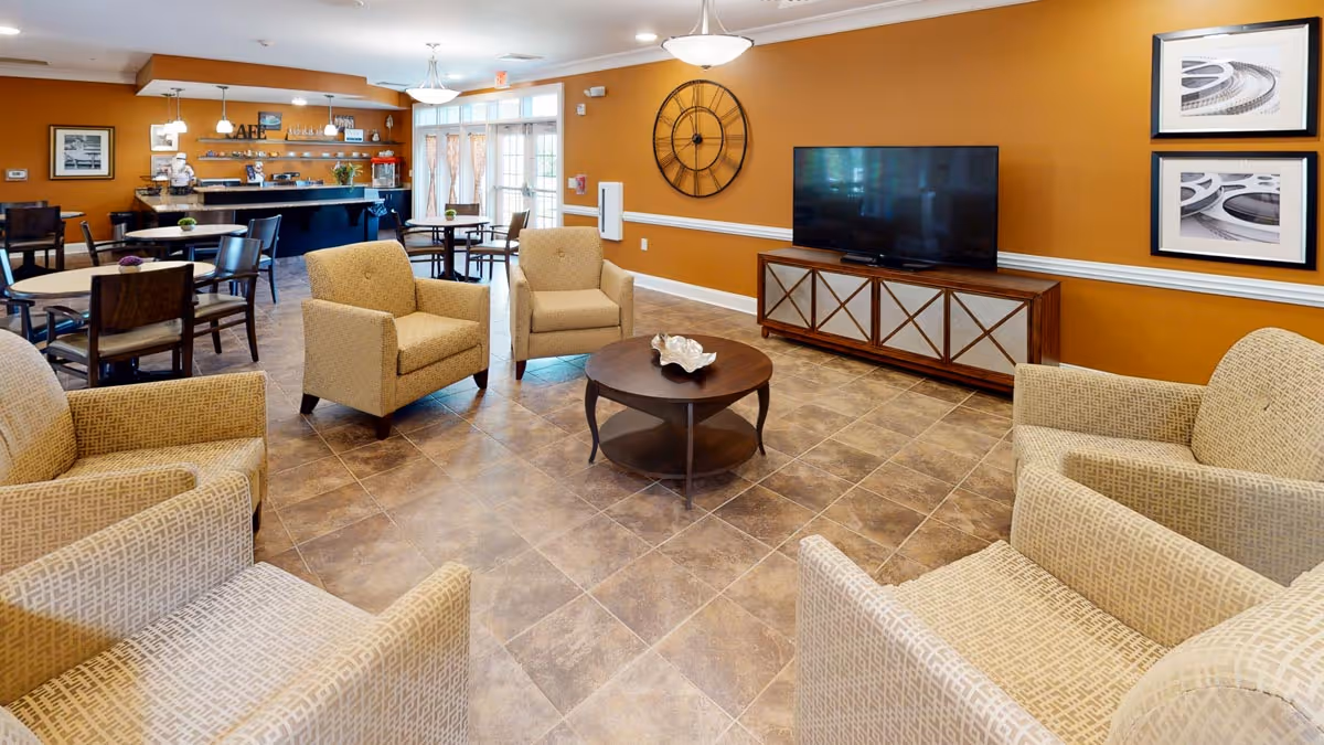 Community lounge with armchairs arranged around a central coffee table, a TV on a console, and dining tables and a serving counter in the background.