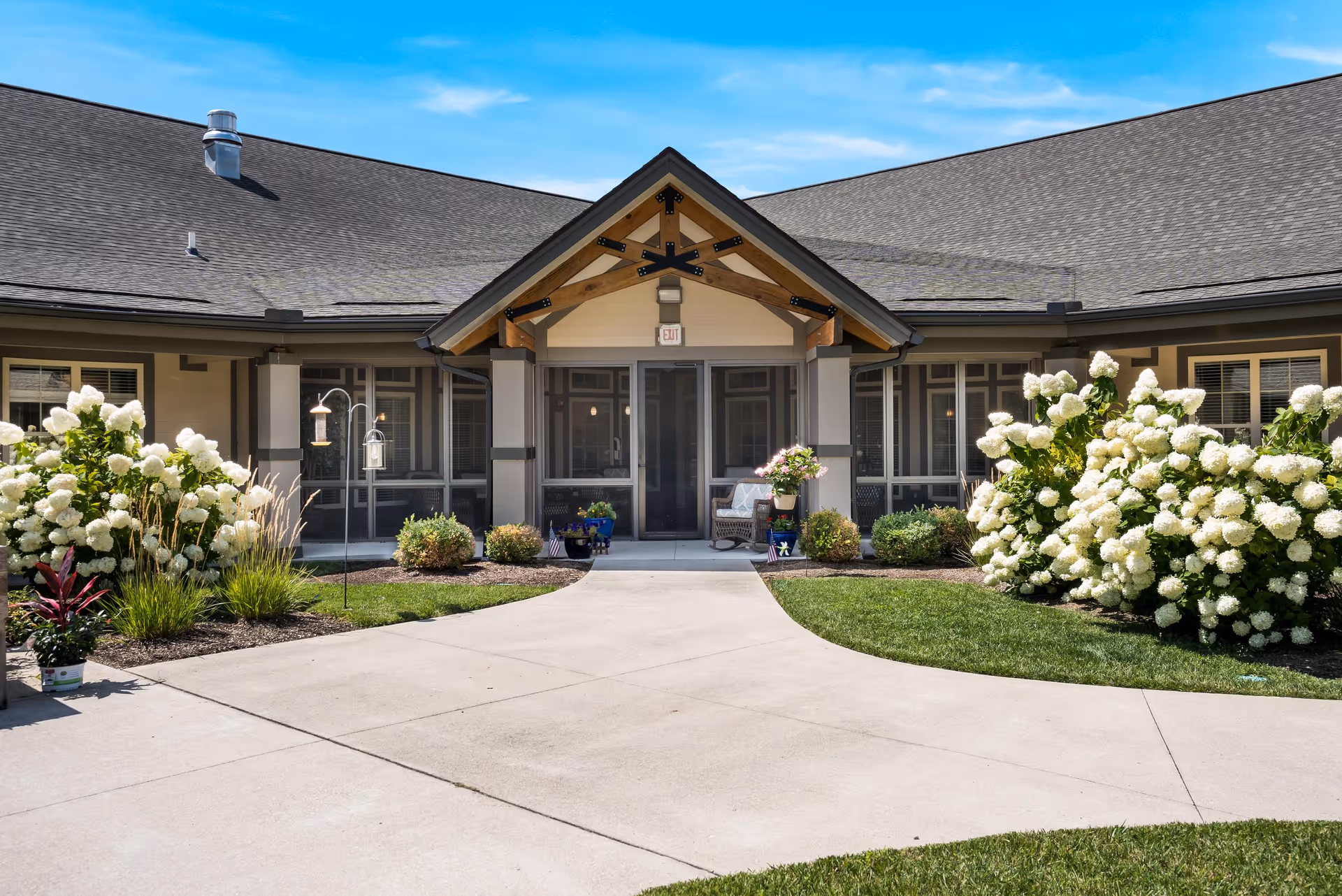 Exterior view of Liberty Place Memory Care facility entrance with a covered porch supported by wooden beams, surrounded by well-maintained landscaping including large white flowering bushes and green shrubs under a clear blue sky.