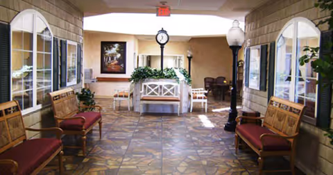 Well-lit senior living facility lobby with benches, potted plants, a central clock and tiled floor.
