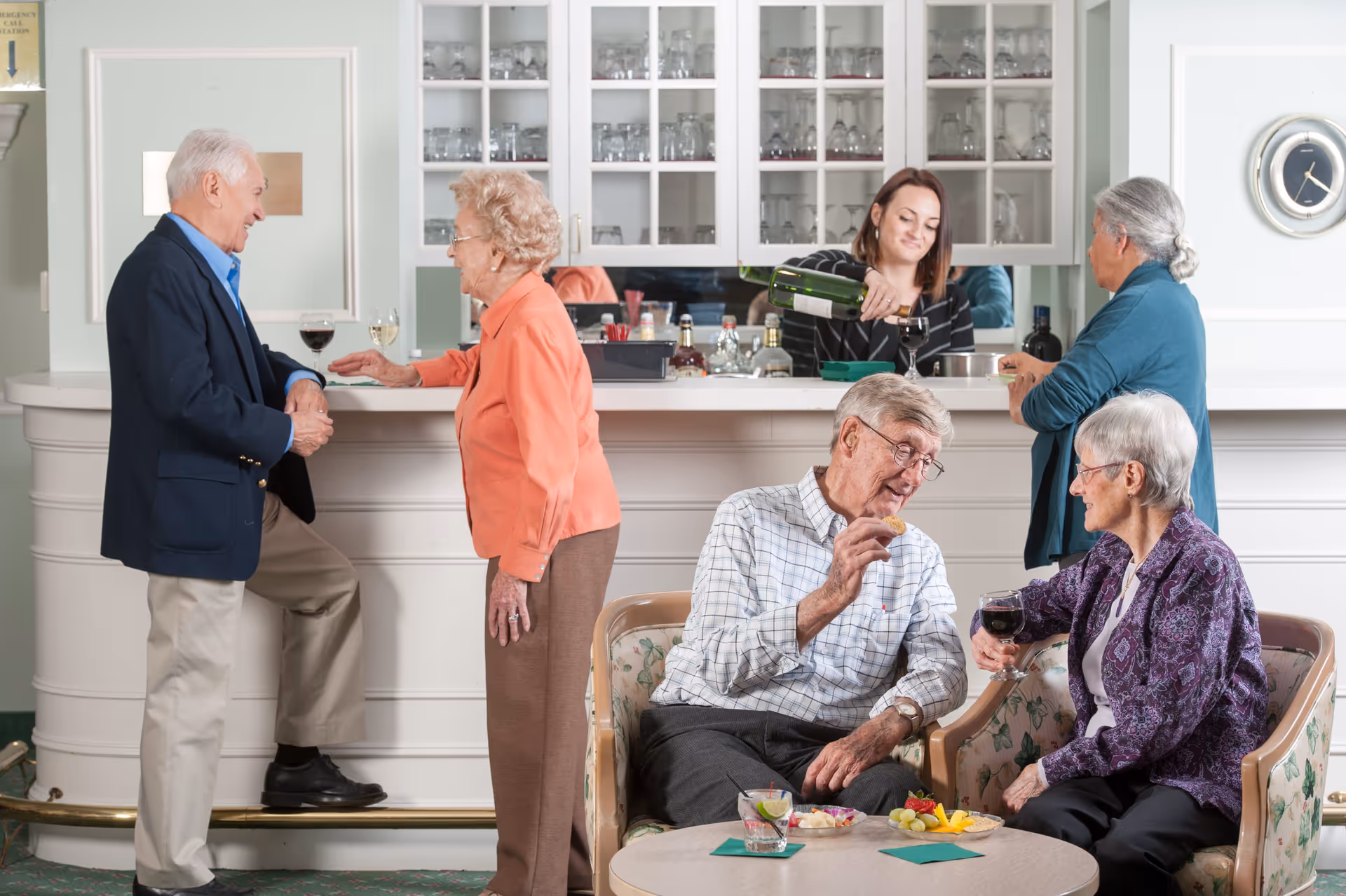 A group of elderly people socializing in a lounge area with a bar. Two elderly men and women are engaged in conversation near the bar counter, where a woman is pouring wine. Another elderly woman stands at the bar, and two elderly people are seated on floral armchairs at a small table with snacks and drinks.