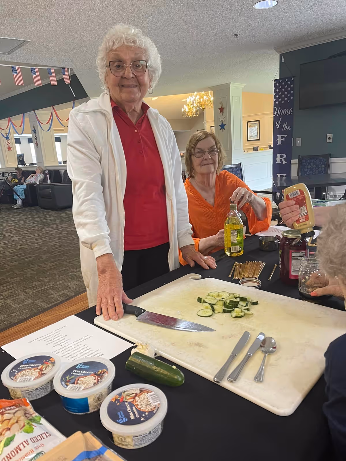 Two elderly women preparing sliced cucumbers on a cutting board in a communal activity/dining area.